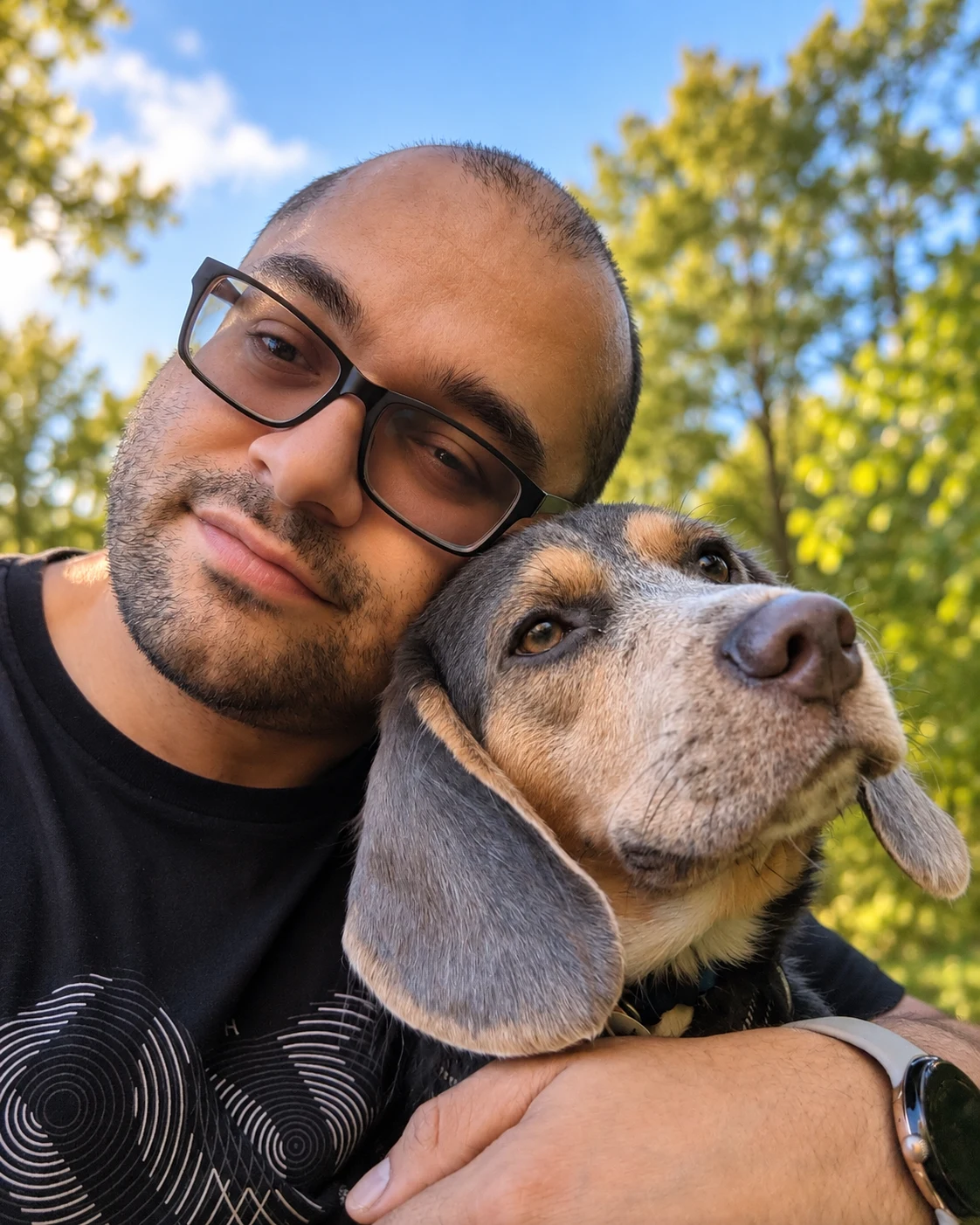 A person taking a selfie outdoors while cuddling Gen, a tricolor beagle, among green trees.